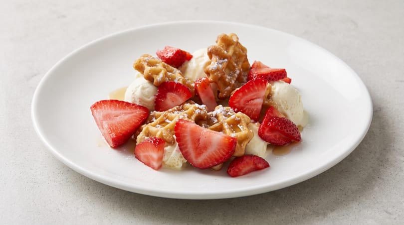 Golden waffles with vanilla ice cream, fresh strawberries, powdered sugar, and syrup on a white plate.
