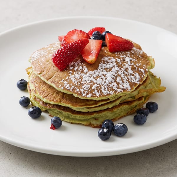Stack of green pancakes with powdered sugar, strawberries, and blueberries on a white plate.
