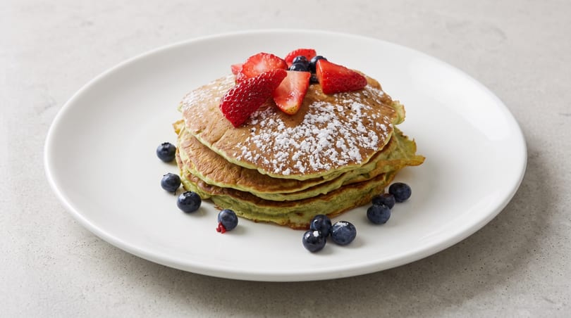 Stack of green pancakes with powdered sugar, strawberries, and blueberries on a white plate.