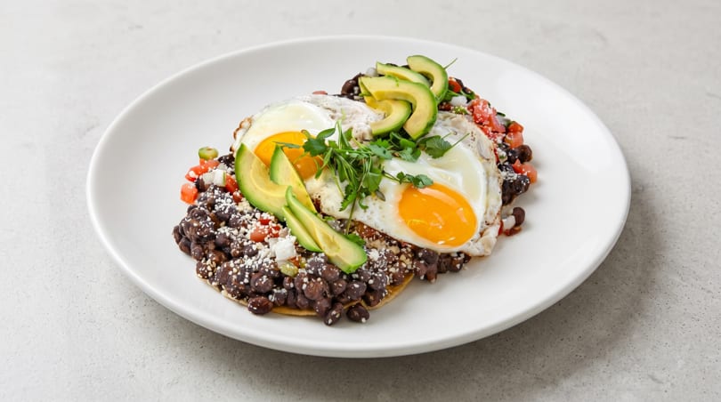Huevos rancheros with two fried eggs, black beans, avocado, and pico de gallo on a white plate.