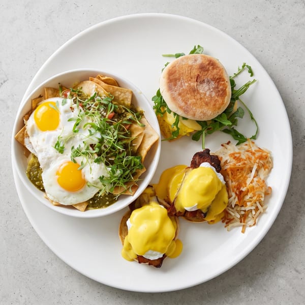 A white plate with chilaquiles, eggs Benedict, hash browns, and a breakfast sandwich.