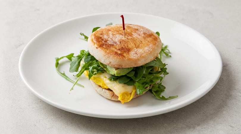 Breakfast sandwich with egg, cheese, avocado, and arugula on an English muffin, served on a white plate.